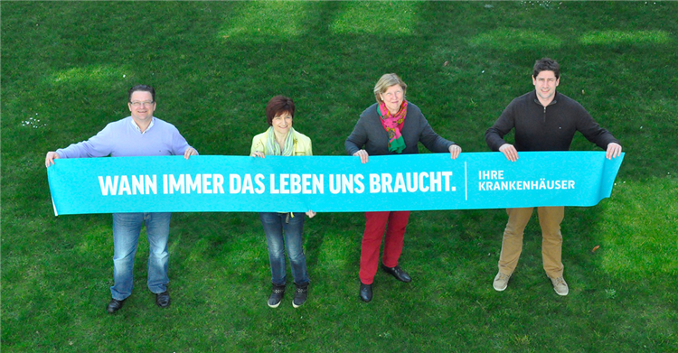 Zwei Männer und zwei Frauen halten auf einer Wiese ein Banner der Kampagne der NRW-Krankenhäuser mit dem Namen Wann immer das Leben uns braucht hoch