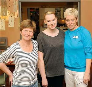 Ein Gruppenfoto mit einer Schülerin und zwei Krankenschwestern, die in einem Wohnraum stehen. Im Hintergrund ist eine Küche zu sehen.