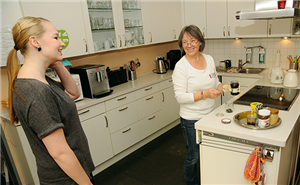 Eine Schülerin und eine Hospiz-Mitarbeiterin stehen lächelnd in einer Küche. Die Hospiz-Mitarbeiterin füllt Marmelade in einen Glasbehälter ab.