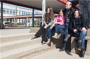 Die beiden Töchter und die Eltern der Familie Demiri sitzen bei starkem Sonnenschein zusammen an bzw. auf einer Mauer. Im Hintergrund ist ein Schulgebäude zu sehen.