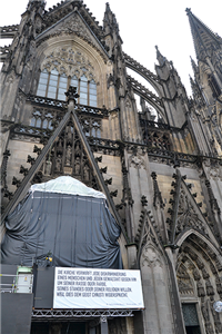 Eine Großaufnahme der Vorderseite des Kölner Doms. An dem, mit einer Leinwand verhängten, Haupteingang befindet sich ein Banner mit einem Auszug aus der 'Nostra Aetate'.