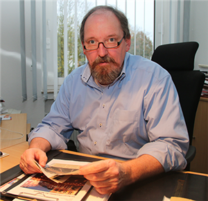 Rolf Leimann sitzt an seinem Schreibtisch vor einem Klemmbrett und hält einen ausgeschnittenen Zeitungsartikel in der Hand.