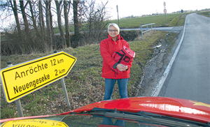 Sandra Rath steht vor ihrem roten Dienstwagen an einer Landstraße. Neben ihr ist ein Schild mit zwei Orts- und Kilometerangaben zu sehen, im Hintergrund erstreckt sich eine Wiesenlandschaft.