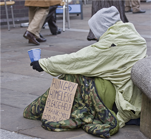 Ein Obdachloser der in einer Fußgängerzone auf dem Boden sitzt und einen Pappbecher hochhält. An seinem Körper lehnt ein Pappschild.