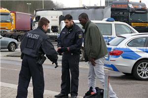 Zwei Polizisten die zusammen mit einem Flüchtling auf einem Parkplatz stehen. Im Hintergrund sind einige Polizeiwagen und LKWs zu sehen.