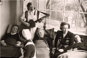 Historische Aufnahme in Schwarz-Weiß: Drei Frauen liegen zugedeckt auf Liegestühlen an einem Fenster. Zwei weitere Frauen sitzen daneben, eine dritte Frau spielt stehend mit einer Gitarre.