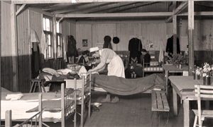 Historische Aufnahme in Schwarz-Weiß: Eine Pflegerin steht in einer Baracke und pflegt eine in einem Krankenbett liegende Person.