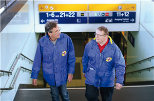 Zwei Mitarbeiter ohne und mit Behinderung von der Bahnhofsmission Essen, die gerade die Treppe zu einem Bahnsteig am Essener Hauptbahnhof hinaufgehen. Im Hintergrund sind Hinweisschilder zu sehen.