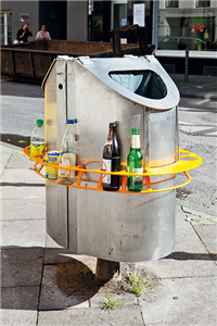 Ein doppelseitiger Mülleimer aus Metall, an dem ein oranger Pfandring befestigt ist, der einige Pfandflaschen enthält. Der Mülleimer steht an einer Straßenecke.