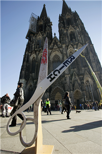 Eine große, geöffnete Schere mit Ständer, die auf der belebten Domplatte mit dem Kölner Dom im Hintergrund. Auf der einen Hälfte der Schere steht der Schriftzug 'Arm', auf der anderen 'Reich'.