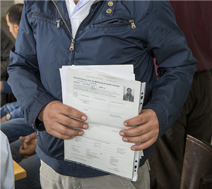 Ein männlicher Flüchtling steht mit andere Flüchtlingen in einem Warteraum und hält einige Dokumente in die Kamera. Der Fokus des Bildes liegt auf den Dokumenten.