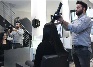 Ein Friseur föhnt einer Frau mit langen, schwarzen Haaren in einem großen Friseursalon die Haare. Beide sind auch im gegenüberliegenden Spiegel zu sehen.