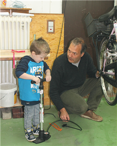 Ein Mann kniet in einem Arbeitsraum neben einem Fahrrad und blickt auf einen Jungen, der links neben ihm an einer großen Luftpumpe steht