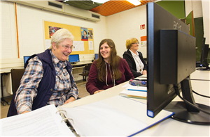 Zwei ältere Frauen und eine Jugendliche sitzen in einem Computer-Raum vor einem Monitor und lachen. Vor dem Monitor liegen einige Schreibutensilien.