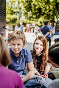 Eine Schülerin und Schüler sitzen mit zwei anderen Kindern auf einem Schulhof einer OGS des Caritasverbandes Lünen zusammen. Im Hintergrund sind andere Schüler und einige Bäume zu sehen.