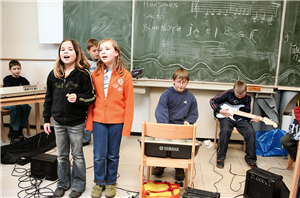 Drei Schüler befinden sich mit verschiedenen Musikinstrumenten in einem Klassenzimmer einer OGS der Hubertusschule in Kevelaer u. mu­si­zie­ren. Davor stehen zwei Mädchen mit einem Mikrofon u. singen.