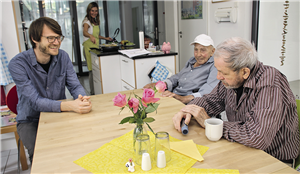 Ein junger Mann und zwei ehemalig obdachlose Senioren und sitzen lachend an einem Esstisch. Im Hintergrund steht eine Frau in einer Küche und kocht.