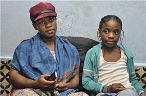 Eine afrikanische Frau, die mit ihrer Tochter auf einem Polster sitzt. Beide wurden frontal fotografiert, die Frau blickt dabei in die Kamera.