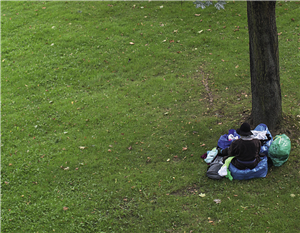 Ein Obdachloser befindet sich auf einer Wiese neben einem Baum und sitzt auf einem blauen Müllsack. Um ihn herum liegen weitere Besitztümer des Obdachlosen.