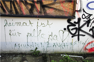 Eine verdreckte, mit Graffiti verschmierte Hauswand, in die ein vernageltes Fenster eingelassen ist. Auf der Wand ist der Spruch 'Heimat ist für alle da' zu lesen.
