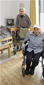 Zwei ehemalig obdachlose Senioren, die sich in einem Wohnraum mit einigen Holzmöbel befinden. Einer sitzt in einem Rollstuhl, der andere hinter ihm steht vor einem Rollator.