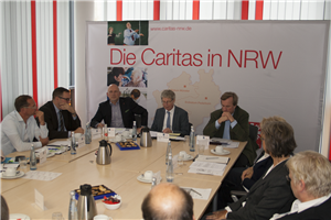Fünf Männer und zwei Frauen sitzen einem Tisch zusammen und diskutieren. Im Hintergrund ist ein Roll-up der Caritas in NRW zu sehen.