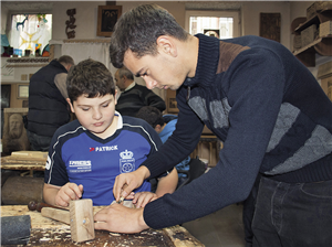 Zwei junge Georgier die zusammen Arbeiten an einem Modell aus Holz verrichten. Im Hintergrund der Werkstatt sind ein weiterer Junge und zwei Männer zu sehen.