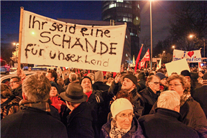Gegendemonstranten einer 'Kögida'-Kundgebung stehen 2015 abends in Köln auf einer Straße und halten Transparente hoch. Auf einem großen Transparent steht: 'Ihr seid eine Schande für unser Land'.