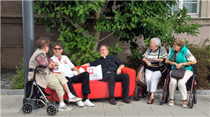 Hubertus Böttcher und Christian Stockmann sitzen auf einem roten Sofa und sprechen mit vier älteren Frauen, die auf ihren Rollatoren Platz genommen haben