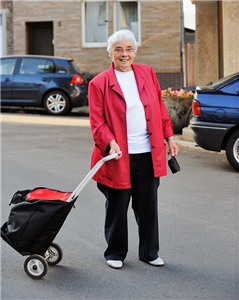 Eine Seniorin steht mit Einkaufskarre auf einer Straße und blickt lächelnd in die Kamera. Im Hintergrund ist ein Wohnhaus und ein schwarzer PKW zu sehen.