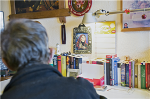 Ein Mann sitzt mit dem Rücken zur Kamera an einem Schreibtisch, auf dem eine große Zahl an Büchern aufgereiht ist. An der Wand hängen unter anderem einige Bilder.