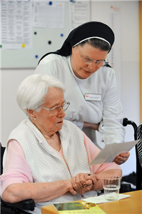 Eine Schwester steht in einem Aufenthaltsraum neben einer Seniorin im Rollstuhl und zeigt ihr ein Schriftstück