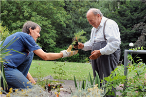 Ein Gärtner kniet in einem Blumenbeet und bekommt von einem Senior eine Pflanze angereicht. Im Hintergrund sind eine Wiese und Bäume zu sehen.
