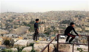 Zwei Jungen befinden sich an einem Berghang nahe der römischen Zitadelle und haben einen Panorama-Blick auf die Stadt Amman. Ein Junge steht am Hang, der andere sitzt auf einem Metallgeländer.