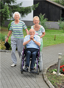 Zwei Seniorinnen und ein Senior im Rollstuhl durchqueren eine Parkanlage über einen Steinweg und lächeln in die Kamera