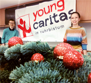 Eine junge Frau und ein junger Mann stehen neben einem Banner der youngcaritas im Ruhrbistum. Im Vordergrund sind Tannenzweige mit roten Weihnachtskugeln zu sehen.
