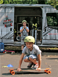 Zwei Jungen mit Sturzhelm befinden sich vor einem Kleinbus. Der eine Junge steht unmittelbar davor, der andere hockt auf einem Skateboard im Vordergrund.