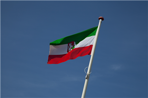 Eine Flagge des Bundeslandes Nordrhein-Westfalen, die an einem weißen Fahnenmast hängt. Im Hintergrund ist der wolkenlose Himmel zu sehen.