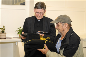 Rainer Maria Kardinal Woelki übergibt einen Mann Küchenmaterial und Decken für den Tagestreff des OCV Leverkusen. Der Mann hält die Decken und Erzbischof Woelki die Küchenartikel in der Hand.