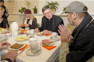Rainer Maria Kardinal Woelki sitzt mit Heinrich Garijs und zwei Frauen an einem gedeckten Frühstückstisch im Tagestreff der Caritas in Leverkusen. Im Hintergrund ist eine weitere Frau zu sehen.