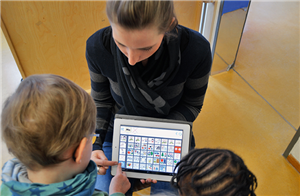 Eine junge Frau kniet mit einem Tablet vor zwei Kindern. Ein Junge bedient eine Talker-App.