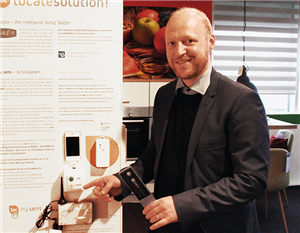 Ein Mann steht in einer Musterwohnung  neben einer Werbewand und deutet auf einige technische Hilfsmittel des Assistenzsystems der Varia GmbH 