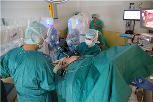 Ein Mediziner und eine Medizinerin hantieren an einem Operationsstisch mit einem OP-Roboter. Hinter einem der OP-Roboter in einer Ecke des Saals haben sich einige Mitarbeiter versammelt.