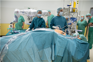 Eine Medizinerin und ein Mediziner stehen an einem Operationstisch. Im Hintergrund sind diverse Geräte und weitere Mitarbeiter zu sehen.