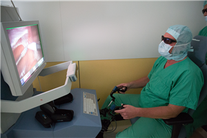 Ein Mediziner sitzt vor dem Steuerungselement eines OP-Roboters und bedient es. Dabei blickt er auf einen Monitor des Roboters.