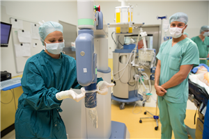 Eine Medizinerin steht neben einem OP-Roboter und stülpt eine Schutzfolie über ihn. Zwei weitere Mediziner stehen daneben.
