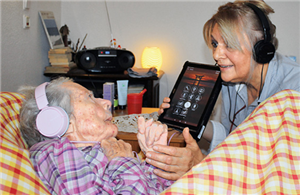 Eine Pflegerin steht mit einem Tablet an einem Bett, in dem eine alte Frau liegt. Beide haben Kopfhörer auf.