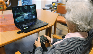 Eine Seniorin sitzt im Gemeinschaftsraum eines Altenheims an einem Tisch vor einem Laptop und spielt ein Videospiel. Sie hält dabei einen XBox-Controller in den Händen.