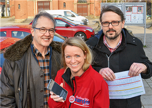 Zwei Männer und eine Frau stehen auf einem Parkplatz, die Frau hält ein Smartphone und einer der Männer ein Dokument in den Händen