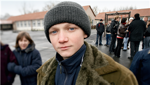 Ein junger Russlanddeutscher blickt in die Kamera. Im Hintergrund stehen auf einem großen Platz viele andere Russlanddeutsche beinander.